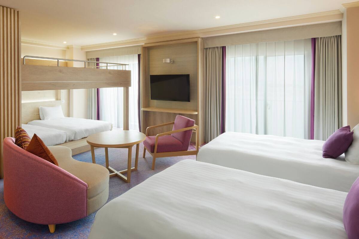 Ein Hotelzimmer mit Stockbetten, einem Schreibtisch, einem Stuhl, einem Fernseher und einem großen Fenster mit Vorhängen.