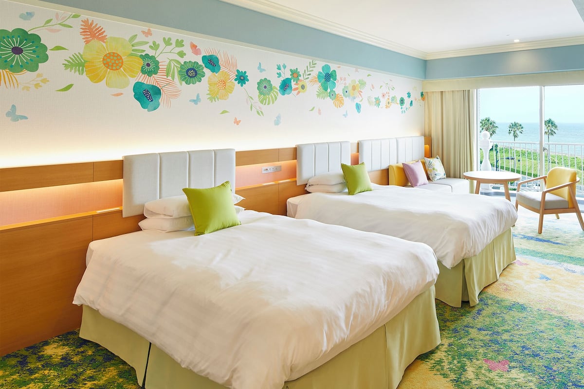 Ein Hotelzimmer mit zwei Betten, einem Fernseher, einem Balkon mit Meerblick und einer Wanddekoration mit Blumenmuster.