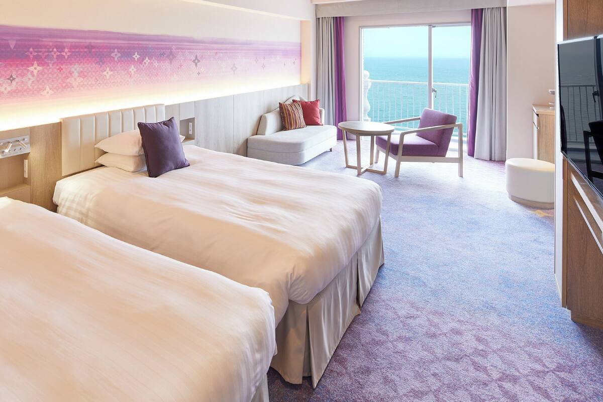 Ein Hotelzimmer mit zwei Betten, einem kleinen Tisch, einem Sessel und einem Fernseher.