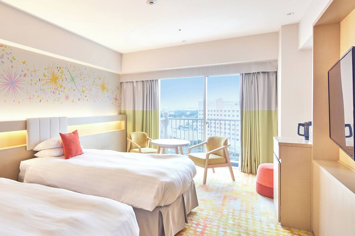 Ein Hotelzimmer mit zwei Betten, einem kleinen runden Tisch und einem Stuhl. Ein großes Fenster bietet einen Blick auf die Stadt.