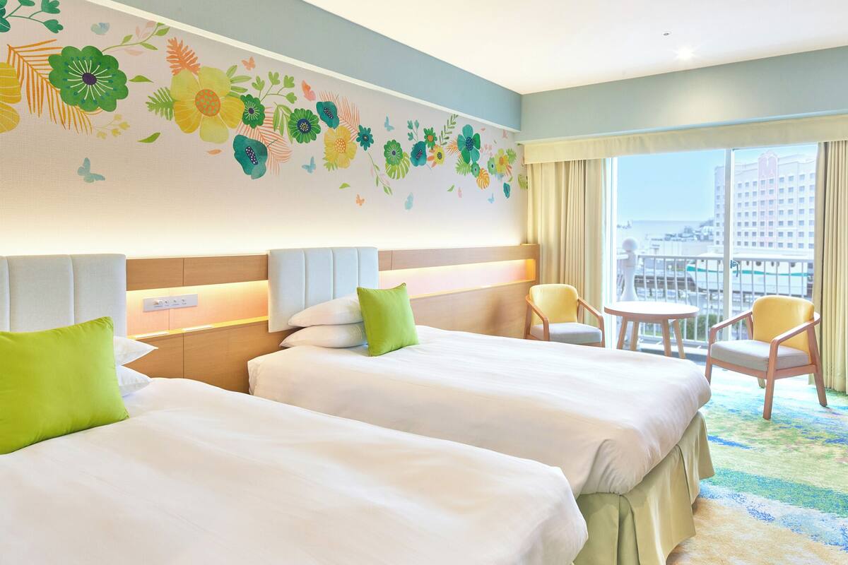 Ein Hotelzimmer mit zwei Betten, einem Schreibtisch, einem Stuhl, einem kleinen Tisch und einem großen Fenster mit Blick auf Gebäude.