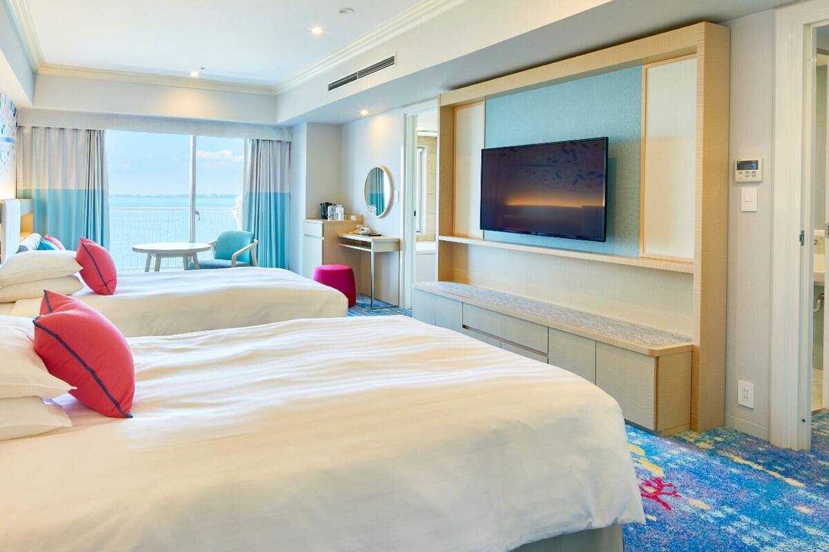 Ein Hotelzimmer mit zwei Betten, einem Fernseher, einem Balkon mit Blick und einem kleinen Tisch.