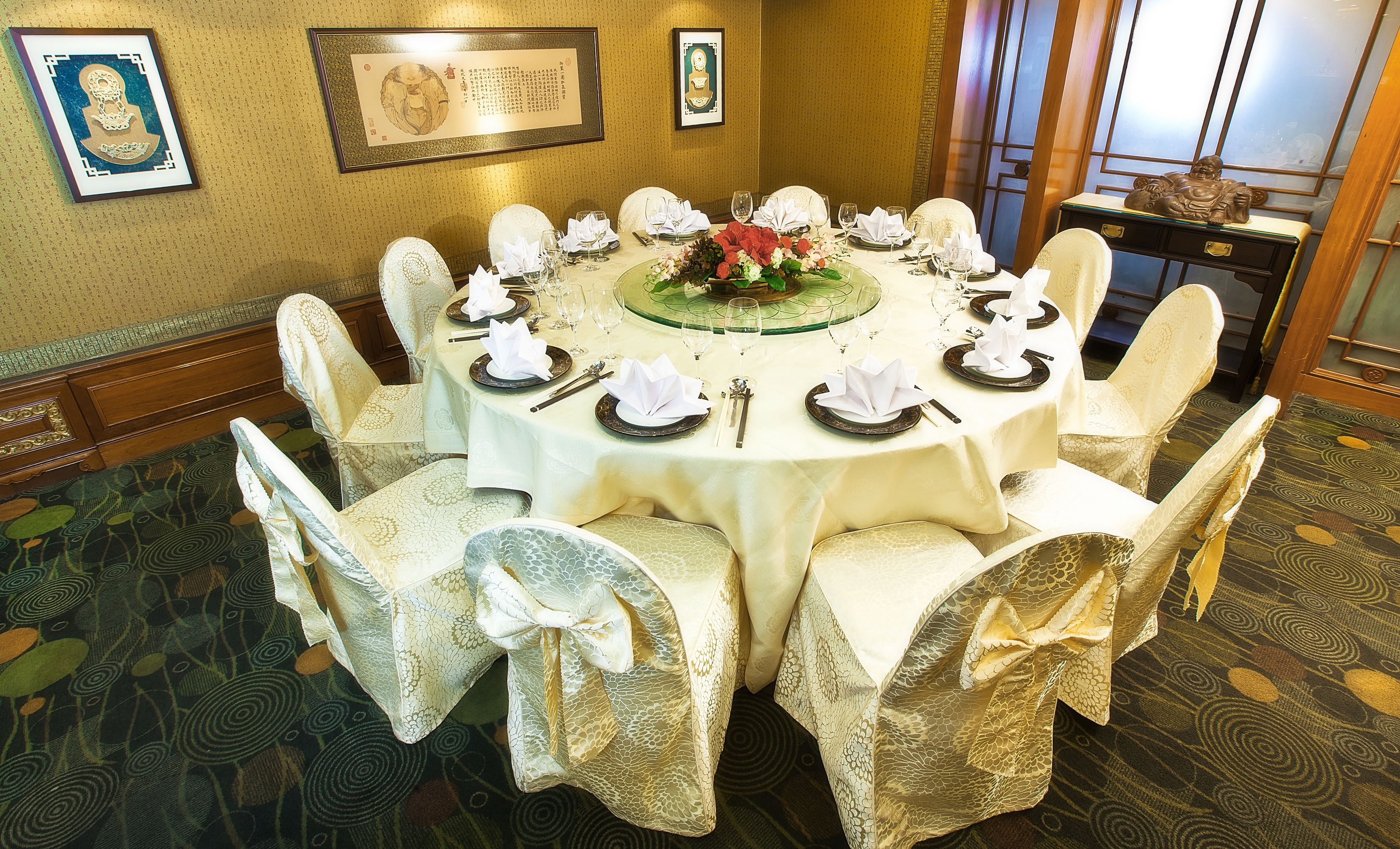 A round dining table set for a formal meal with white chairs, a floral centerpiece, and framed artwork on the walls.