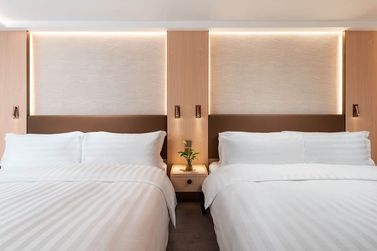 A hotel room with two beds, a nightstand with a lamp, and a wooden headboard with built-in lighting.