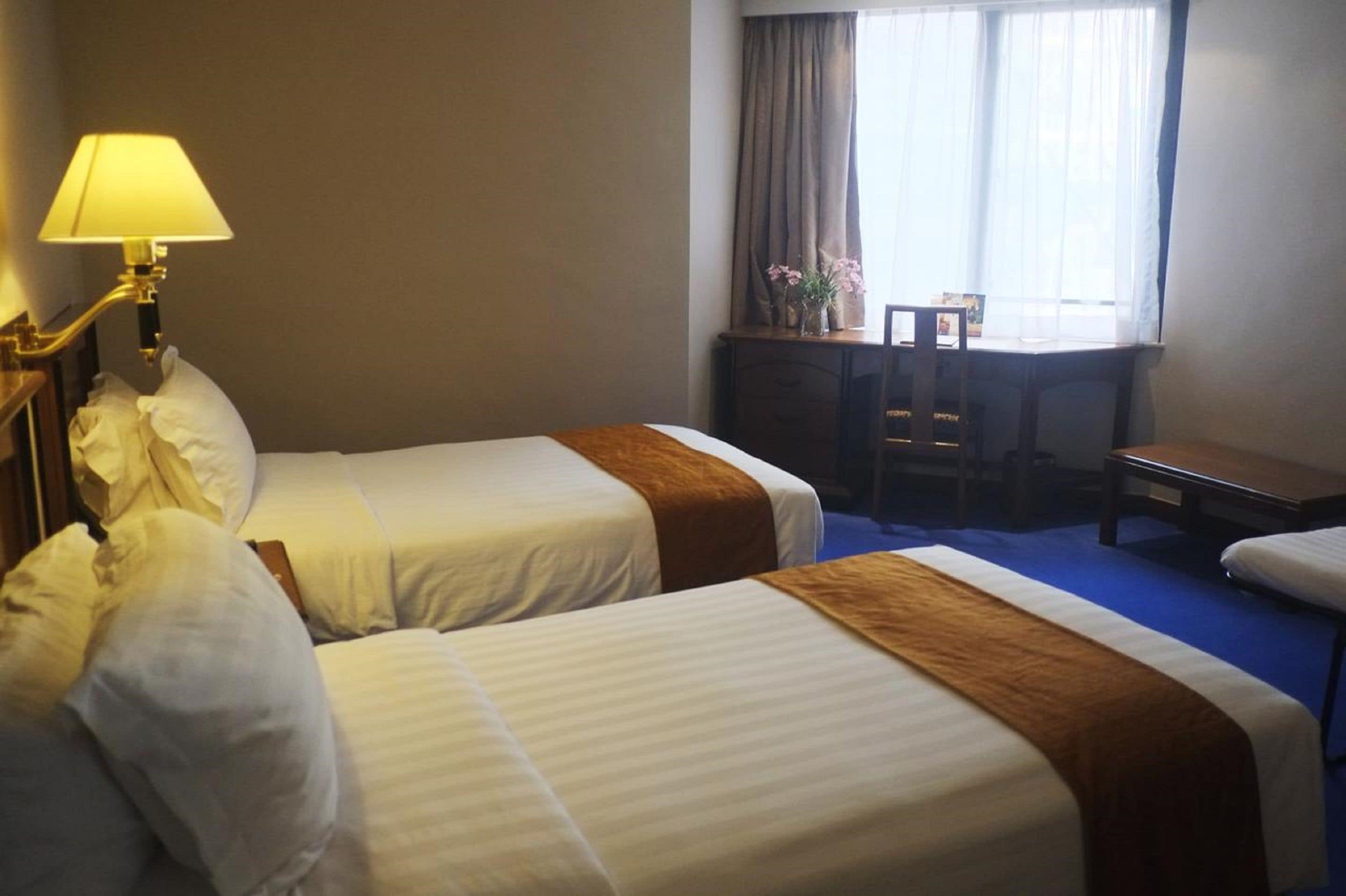 A hotel room with two beds, a desk, a chair, and a lamp.