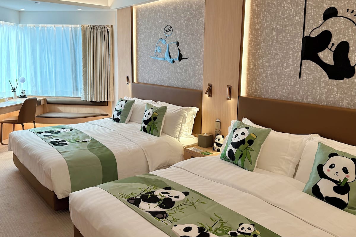 A hotel room with two beds, each with panda-themed bedding and pillows. There is a desk with a chair, a window with curtains, and wall art depicting pandas.