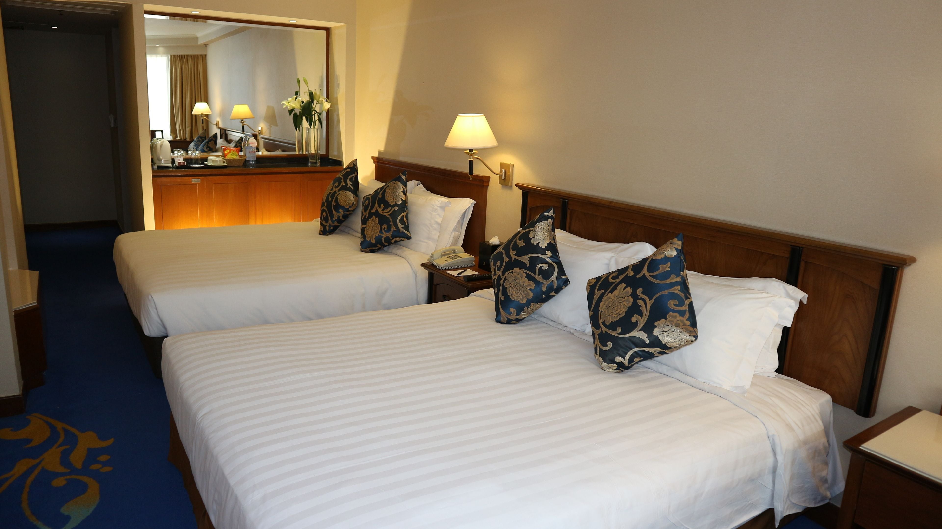 A hotel room with two beds, each with white linens and decorative pillows, a bedside table with a lamp, and a mirror reflecting the room.