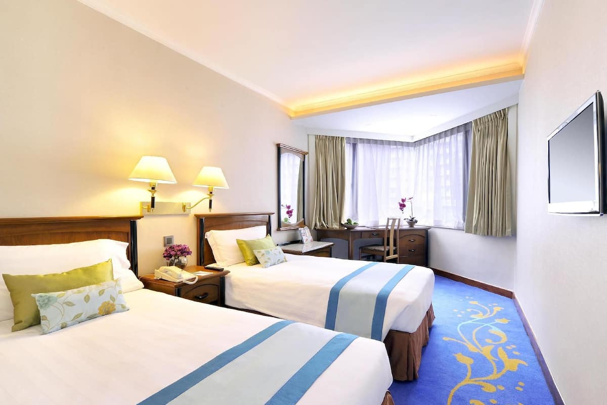 A hotel room with two beds, a desk, a chair, a lamp, a mirror, and a window with curtains.