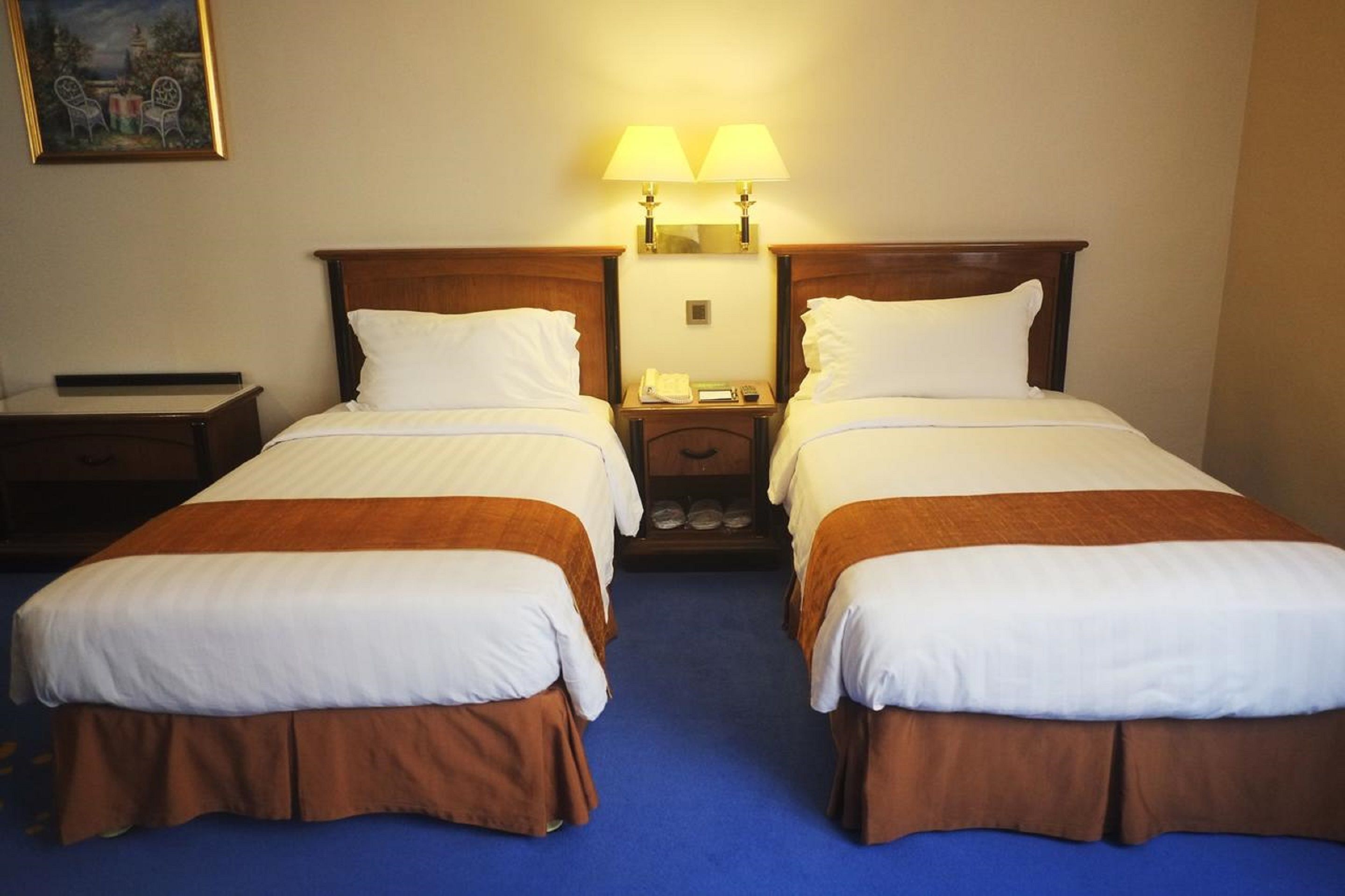 A hotel room with two single beds, a nightstand, a lamp, and a painting on the wall.