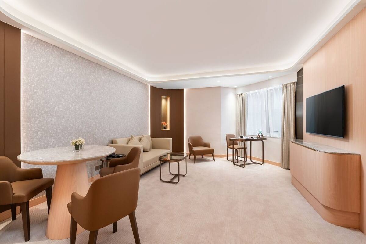 A modern hotel room with a round dining table, sofa, armchairs, a TV mounted on the wall, and a large window with curtains.