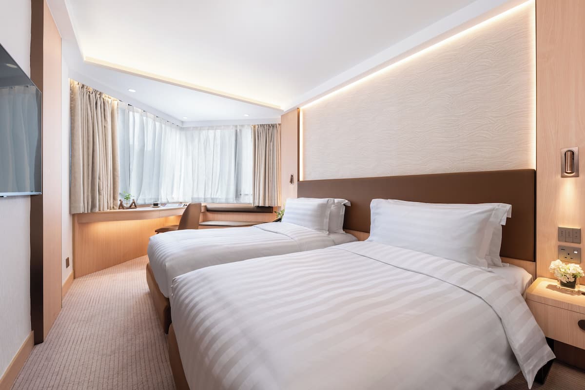A hotel room with two beds, a window with curtains, a bedside table with a vase of flowers, and a wall-mounted light fixture.