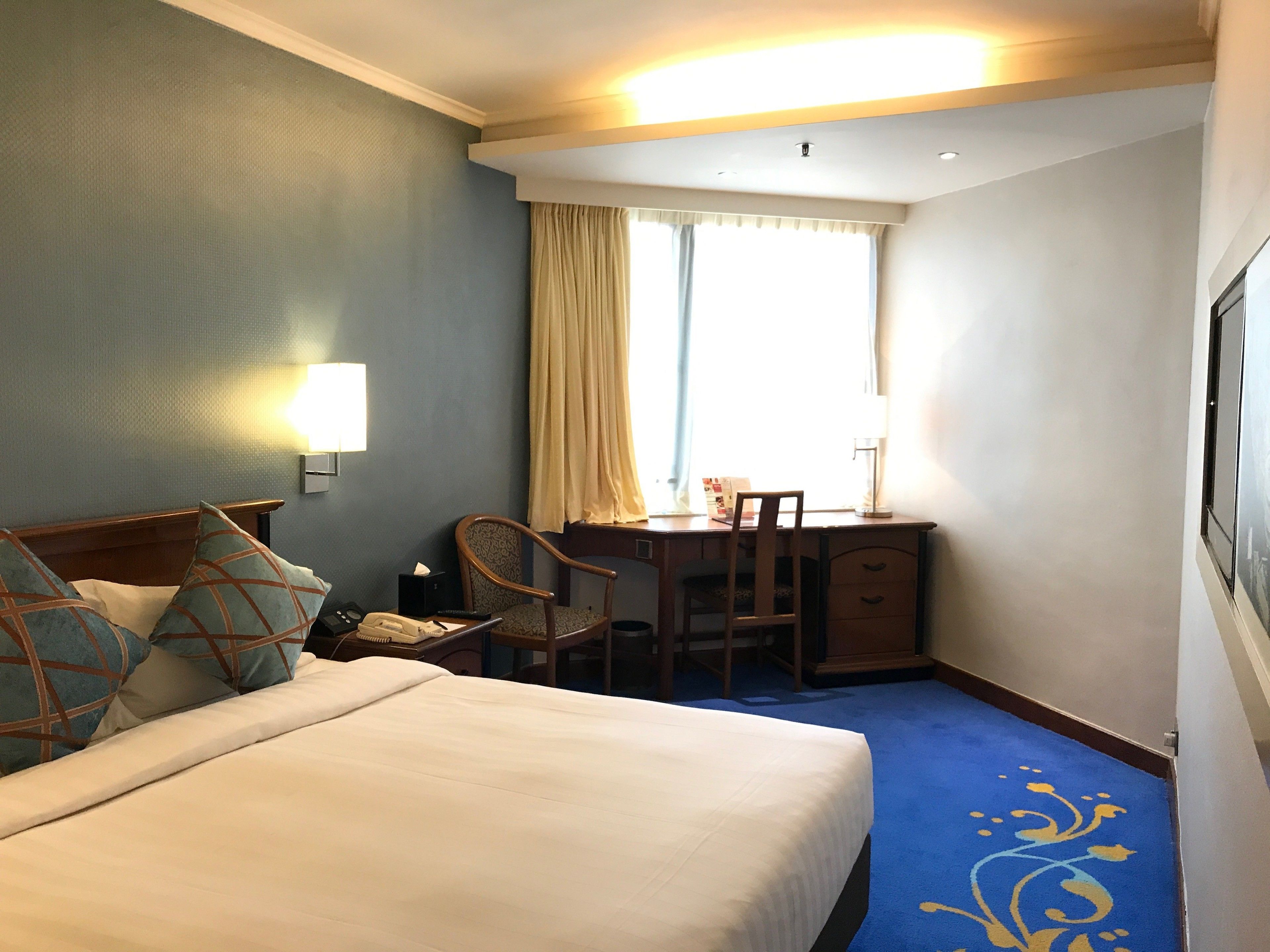 A hotel room with a large bed, a desk with a chair, a lamp, a window with curtains, and a wall-mounted phone.