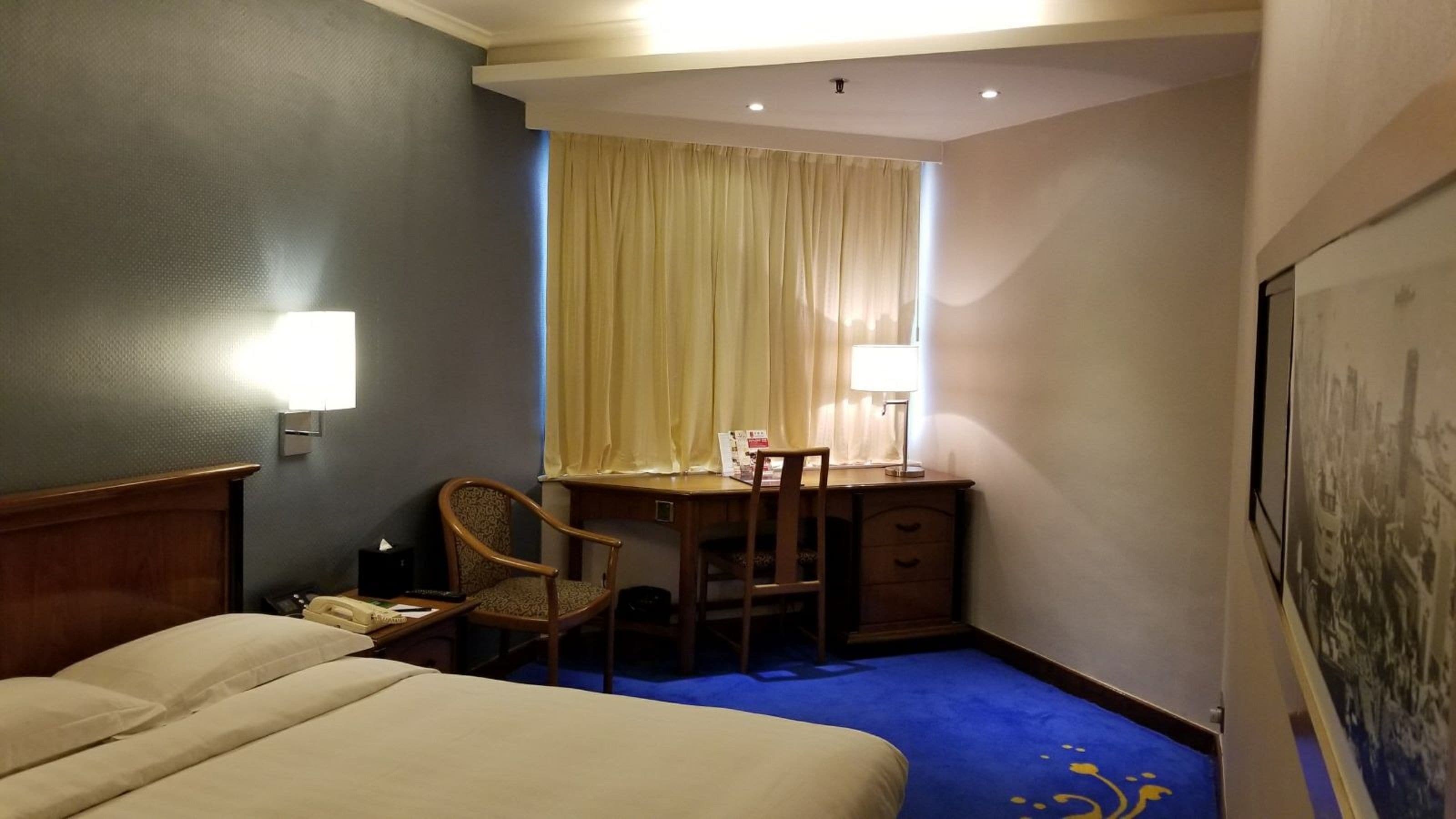 A hotel room with a bed, a desk, a chair, a lamp, and a window with curtains.