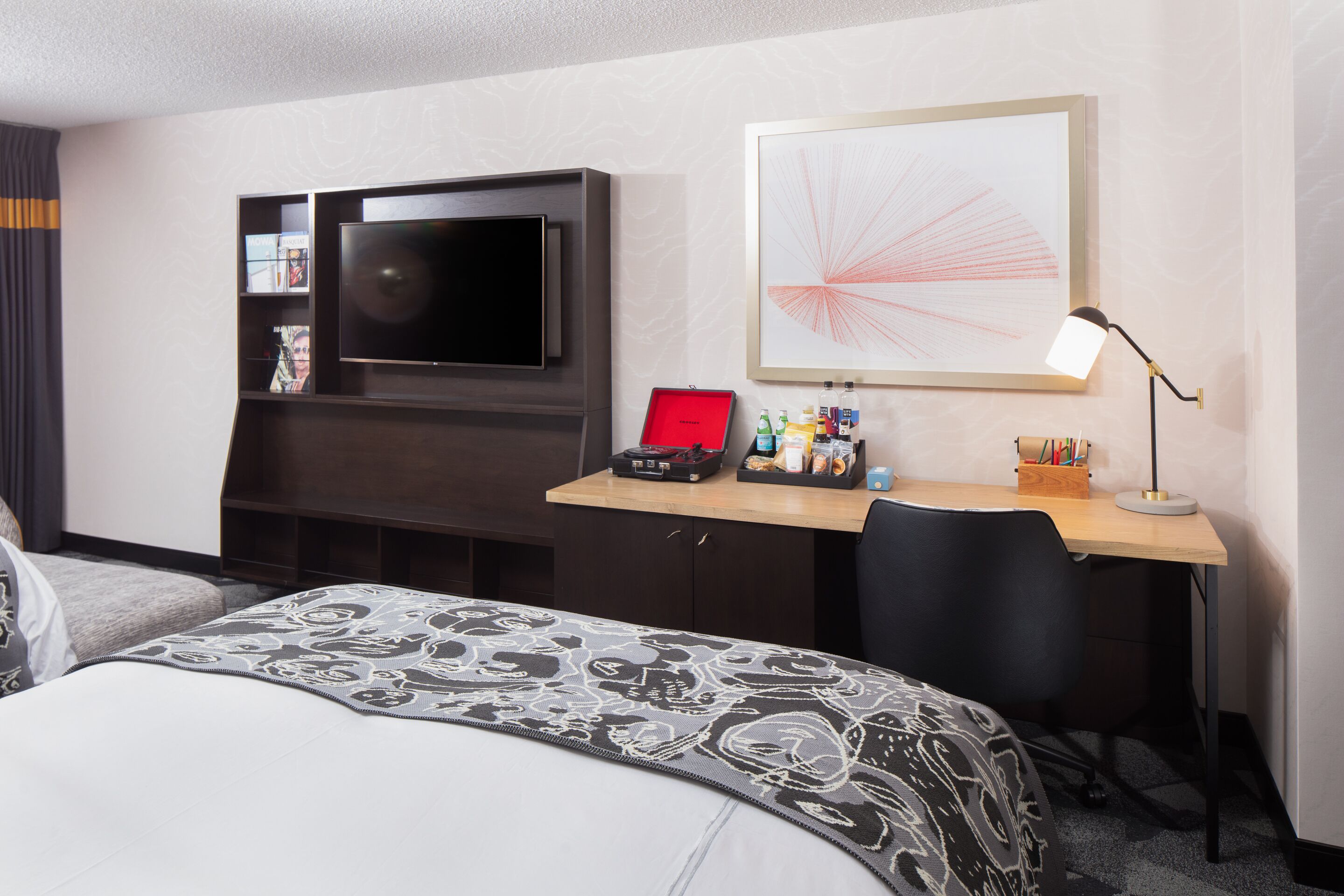 Quarto de hotel com uma cama, uma escrivaninha com computador, televisão e uma obra de arte emoldurada na parede.