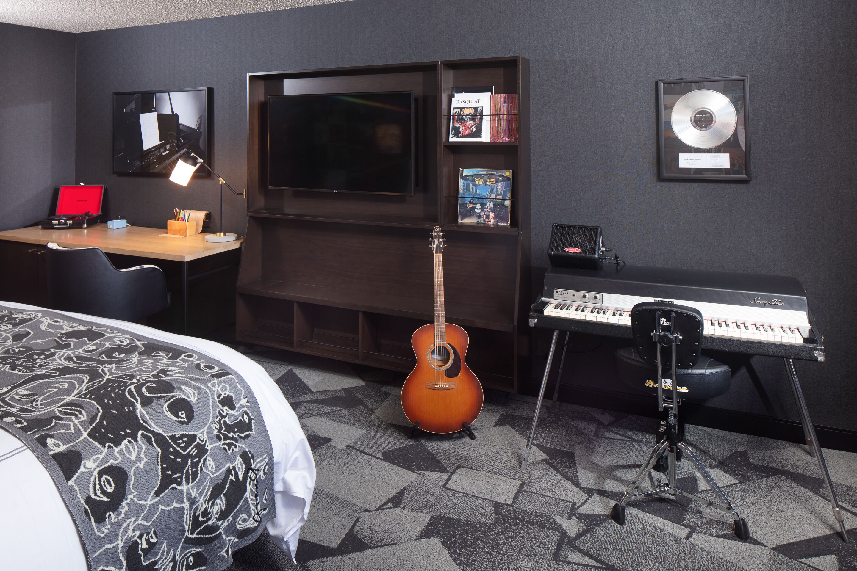 A hotel room with a bed, a desk with a computer, a guitar, a keyboard, a TV, and framed awards on the wall.