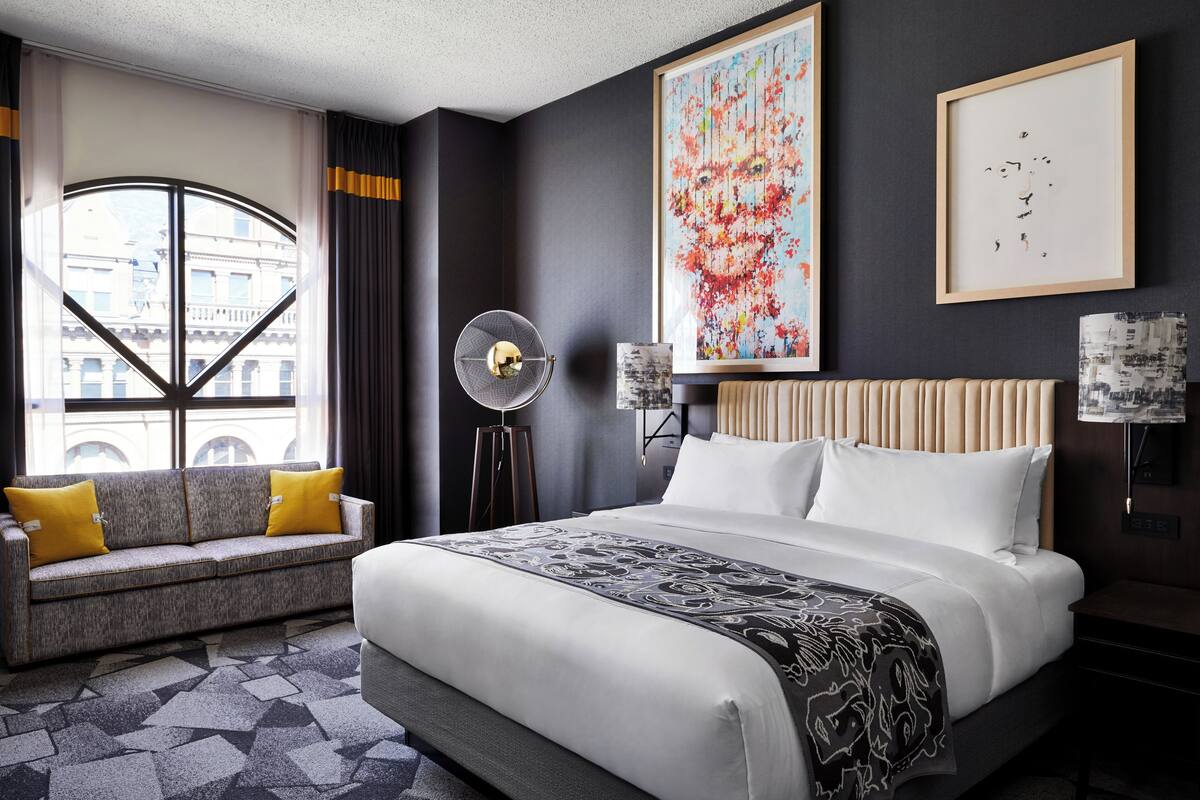 Une chambre d’hôtel moderne avec un grand lit, un canapé avec des coussins jaunes, un ventilateur sur pied et des œuvres d’art accrochées aux murs.