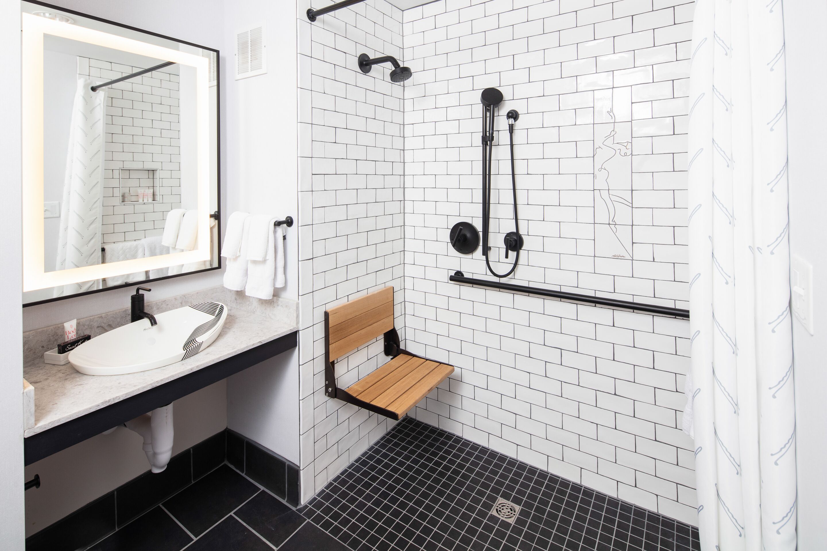 A modern bathroom with a white-tiled shower, a wooden bench, a black sink, and a mirror with a lighted frame.