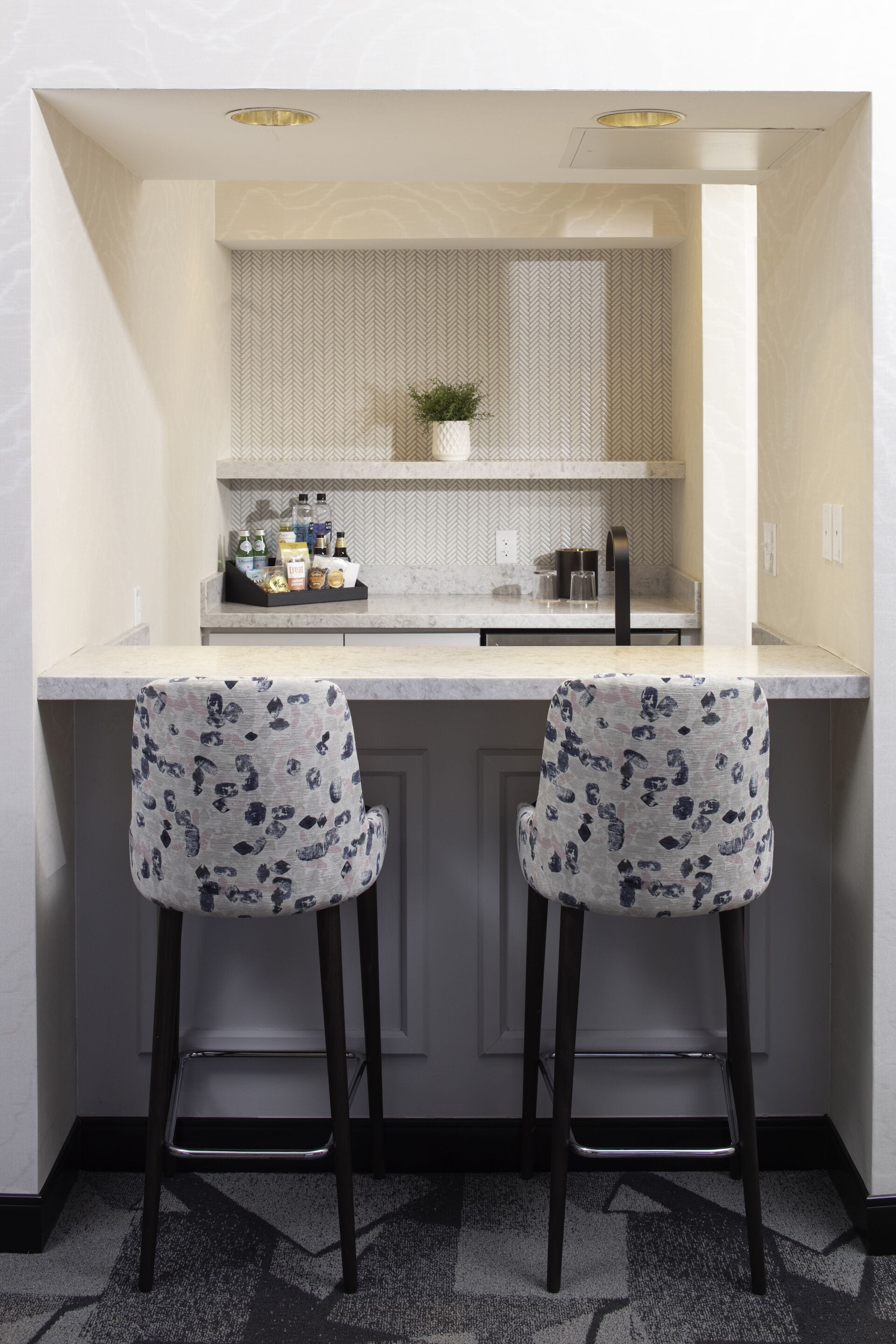 A kitchen area with two bar stools, a countertop with a herringbone backsplash, and a small potted plant.