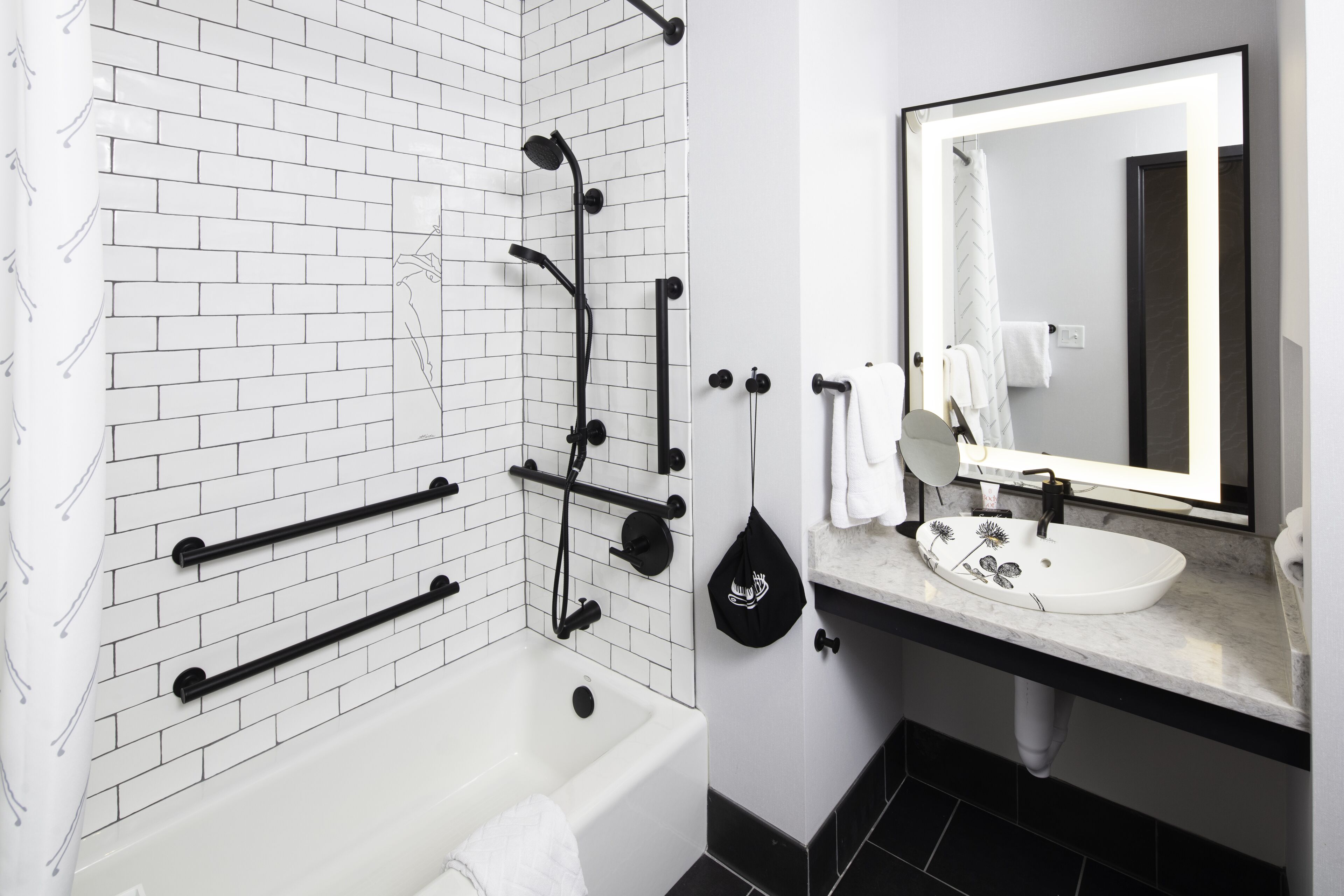 Un baño moderno con ducha de azulejos blancos, toallero negro, lavabo blanco con espejo y suelo negro.