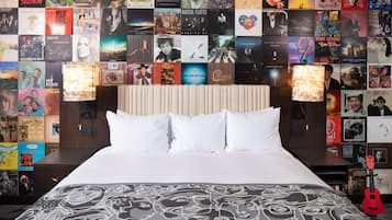 A hotel room with a large bed, a headboard with posters, and a guitar on the nightstand.