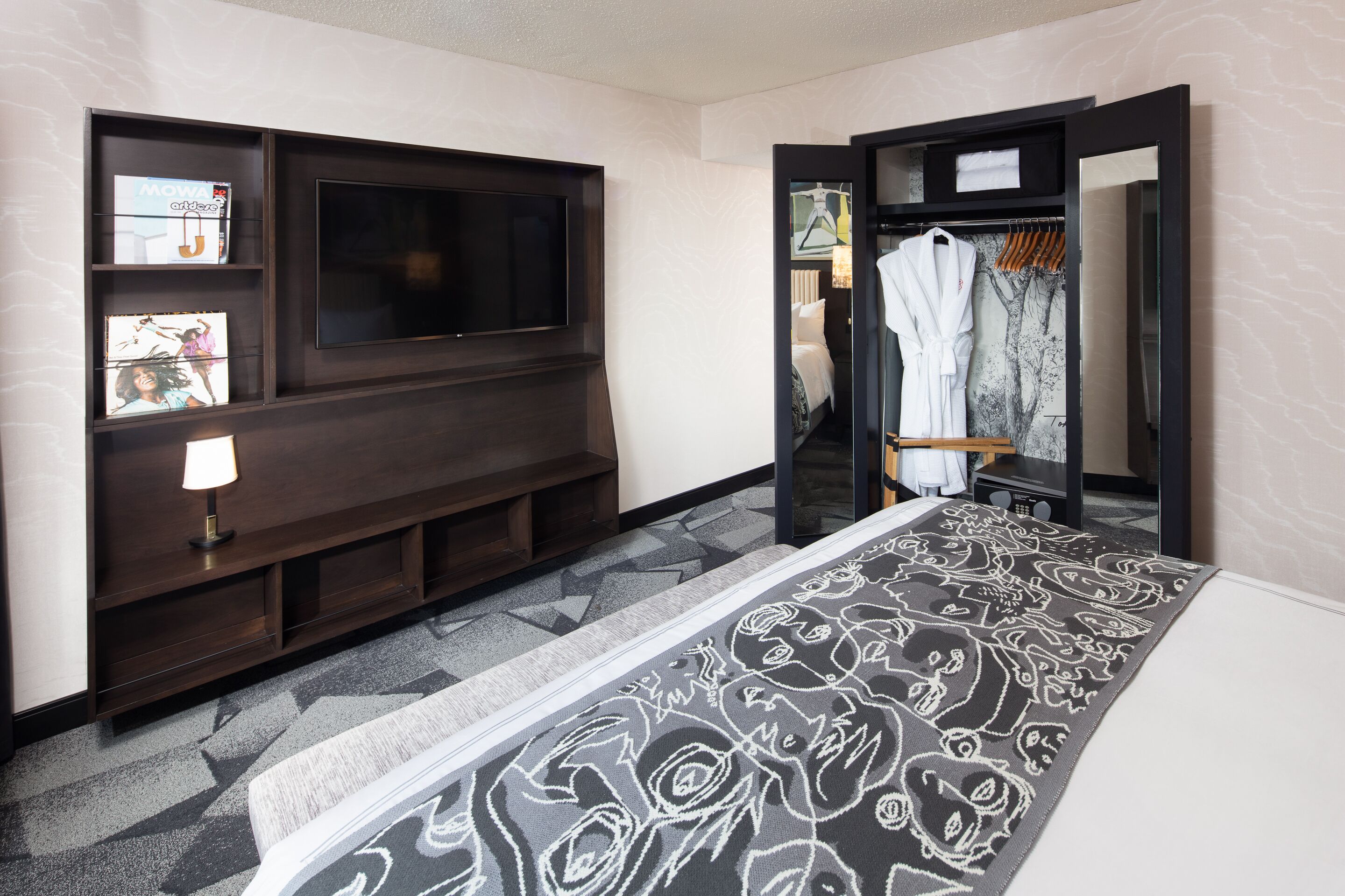 A hotel room with a large bed, a flat-screen TV mounted on the wall, a wardrobe with a mirror, and a small seating area with a lamp.