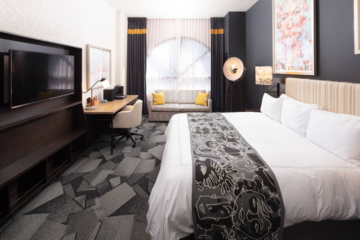Une chambre d’hôtel avec un grand lit, un bureau avec une chaise, une télévision, un canapé avec des coussins, une lampe sur pied et une grande fenêtre avec des rideaux.