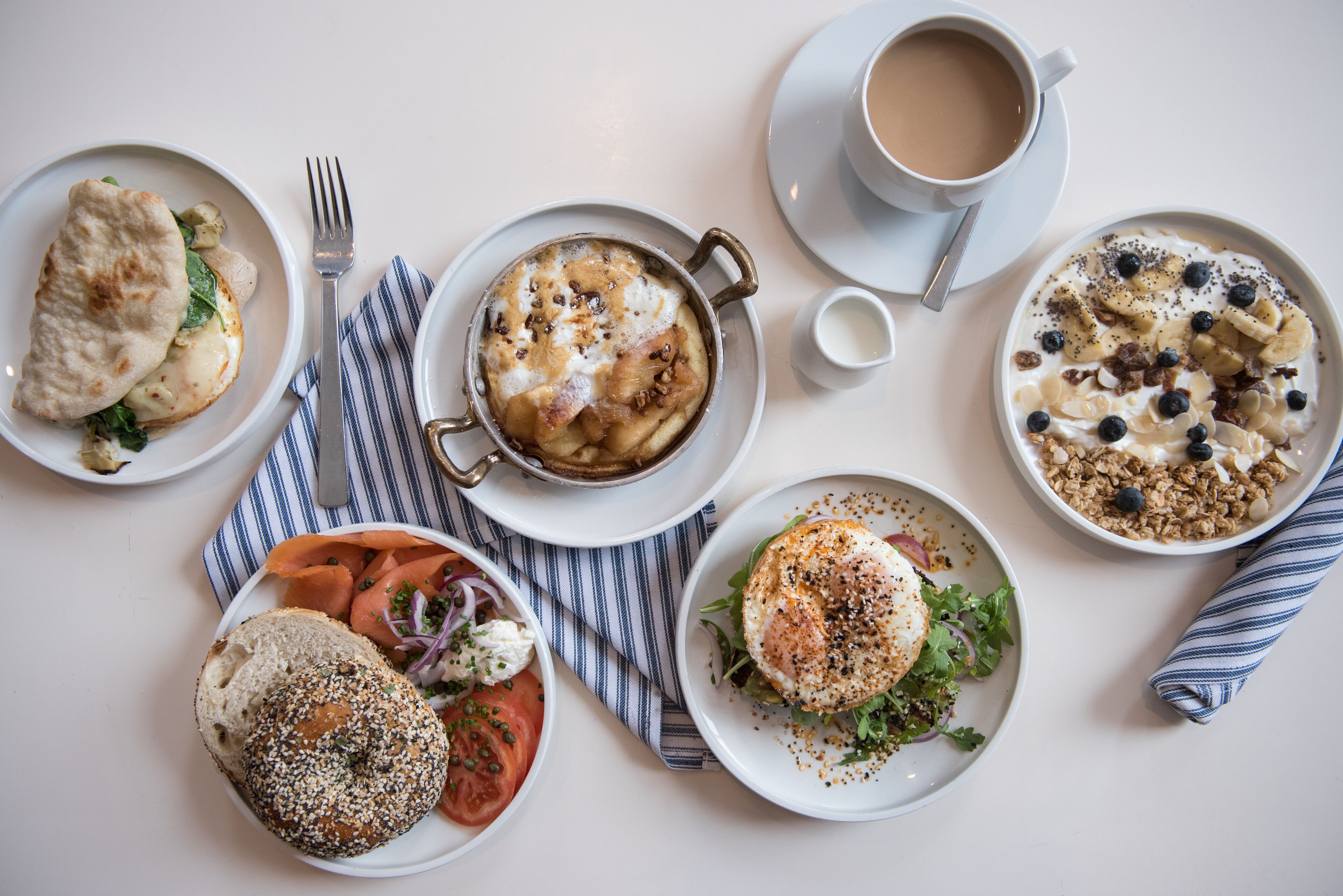A breakfast setting with various dishes, including a bagel, omelette, and yogurt parfait, on a white table.