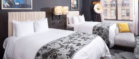 A hotel room with two beds, a nightstand, a lamp, and a view of the city through the window.