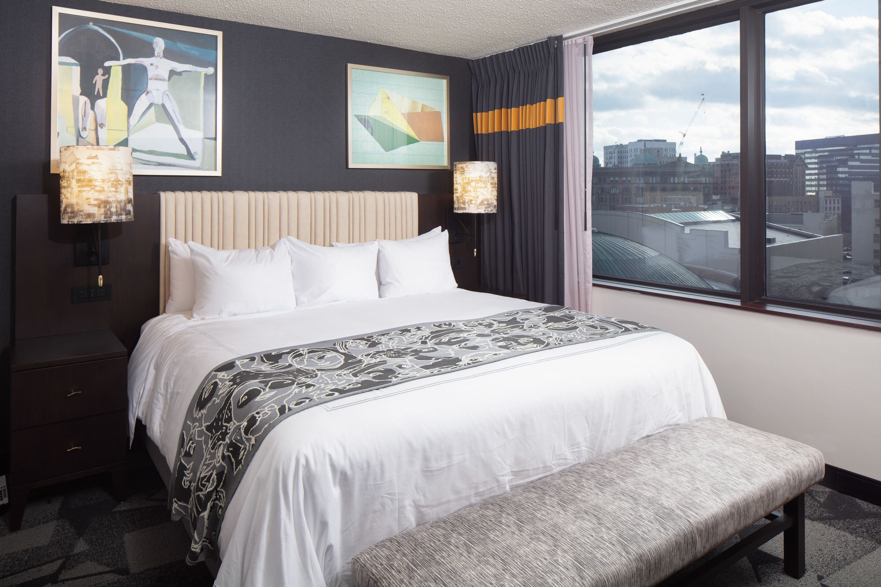 A hotel room with a large bed, a bench, two wall-mounted lamps, and a city view through a large window.