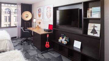 A hotel room with a large TV, a desk with a laptop, a guitar, and a bookshelf.