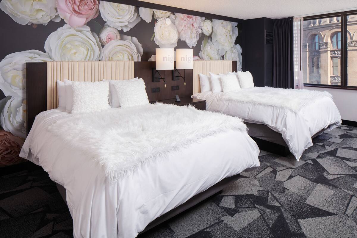 Une chambre d’hôtel avec deux lits, une grande fresque murale à motifs floraux et une vue sur un immeuble par la fenêtre.