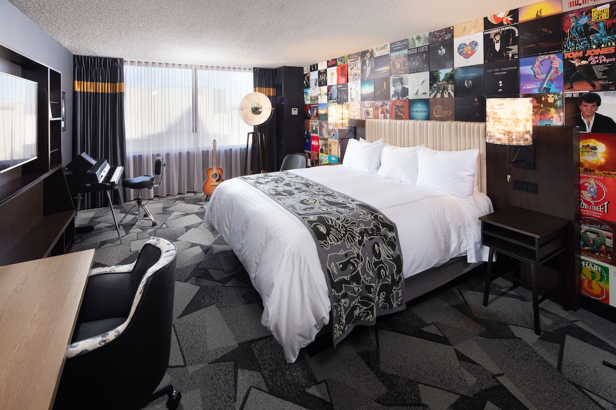 Une chambre d’hôtel avec un grand lit, un bureau avec un ordinateur, une chaise, une guitare et un mur décoré d’affiches de films.