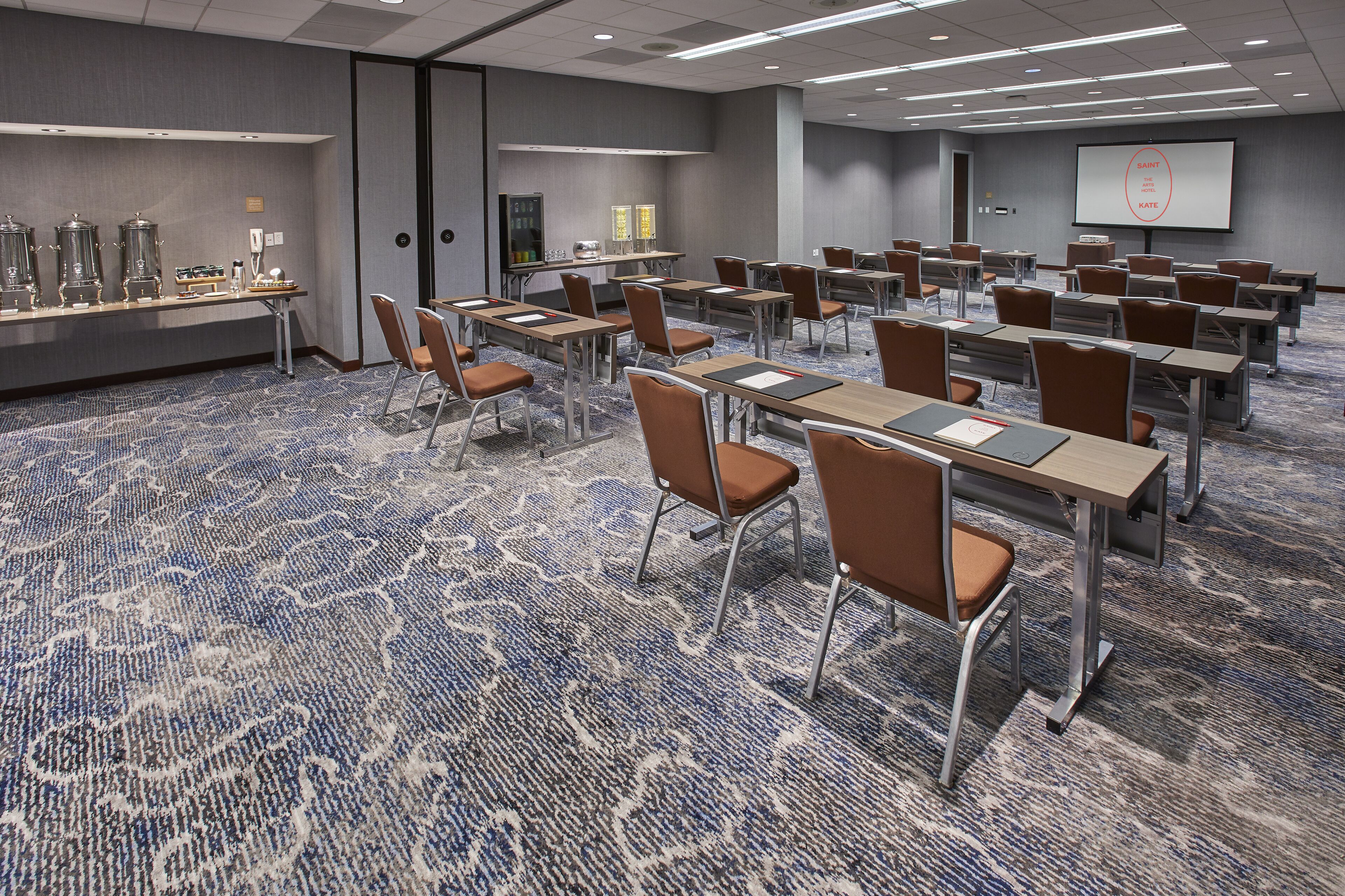 A conference room with rows of tables and chairs, a projector screen, and a coffee station.