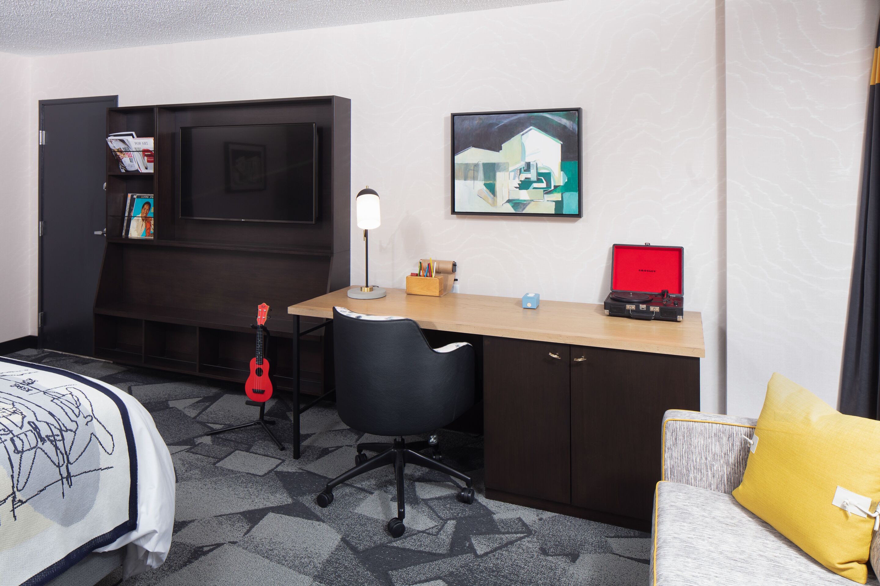 Et hotelværelse med en seng, et skrivebord med en computer, en stol, en guitar og et billede på væggen.