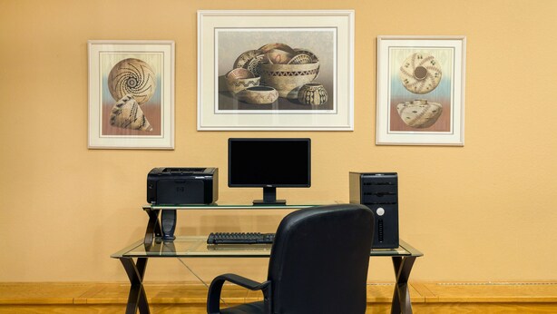 A modern office desk with a computer, printer, and chair, set against a beige wall with framed artwork above. - Days Inn by Wyndham Taos (Taos)
