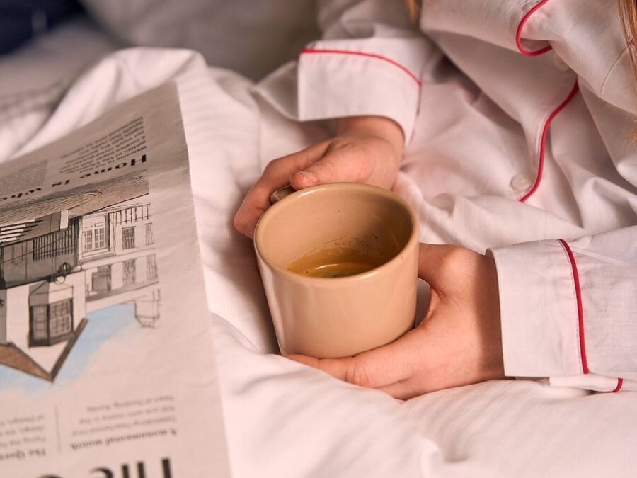 A person in white pajamas with red trim, holding a beige mug, sitting on a bed with a newspaper featuring a building illustration.