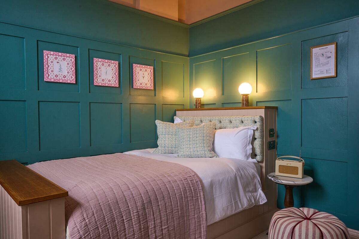 A teal-colored bedroom with a bed, bedside lamps, a small table with a radio, and decorative wall panels.