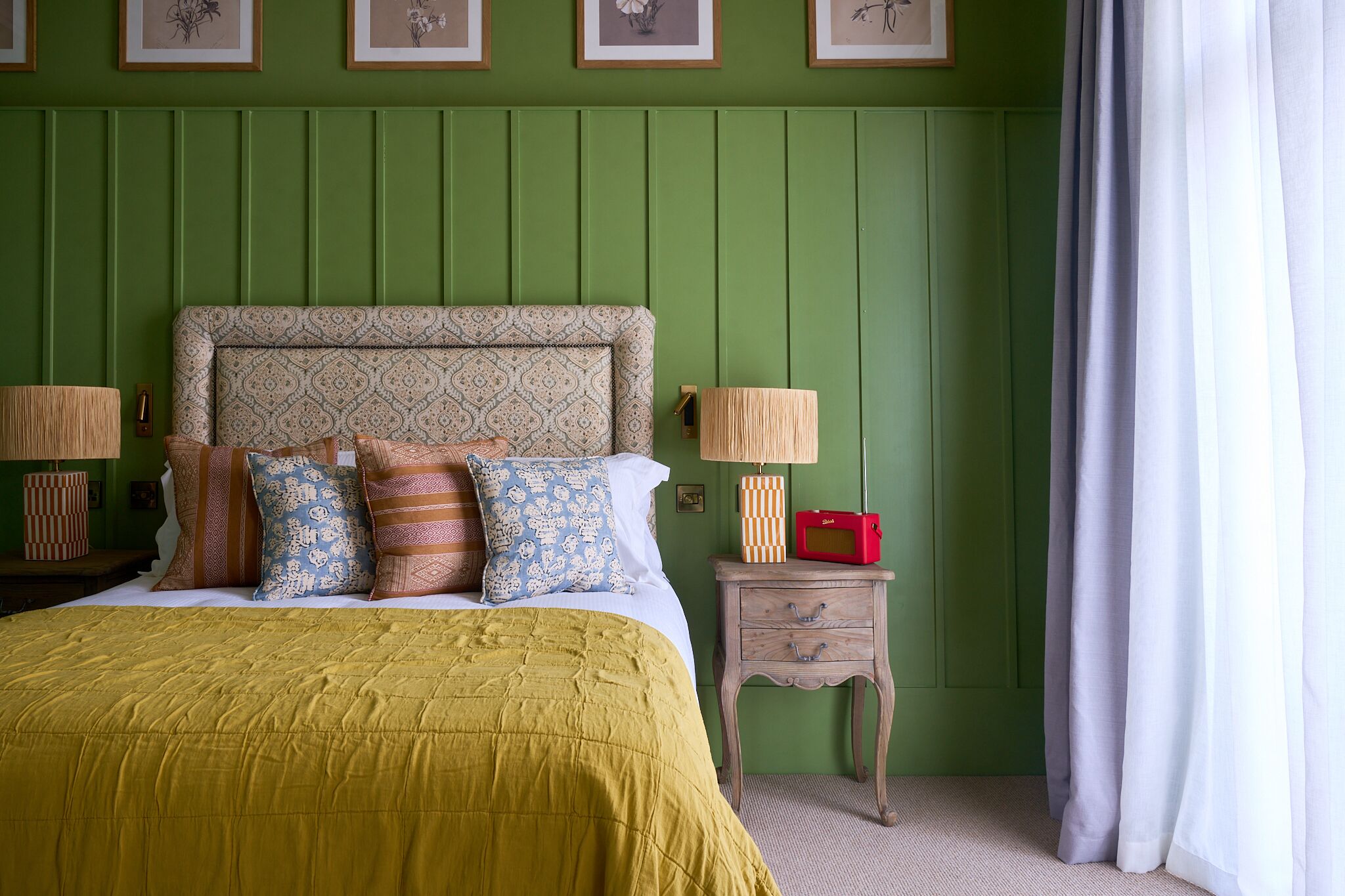 Quarto com parede revestida de verde, cabeceira estampada, cama com várias almofadas, abajures, mesa de cabeceira de madeira pequena e um rádio vermelho.