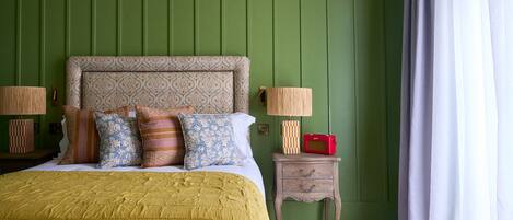 A bedroom with a green paneled wall, a patterned headboard, a bed with multiple pillows, bedside lamps, a small wooden nightstand, and a red radio.