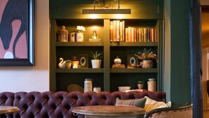 A cozy seating area with a tufted sofa and a patterned armchair, a small round table, and a bookshelf with decorative items. - The White Horse - A Heartwood Inn (Dorking)