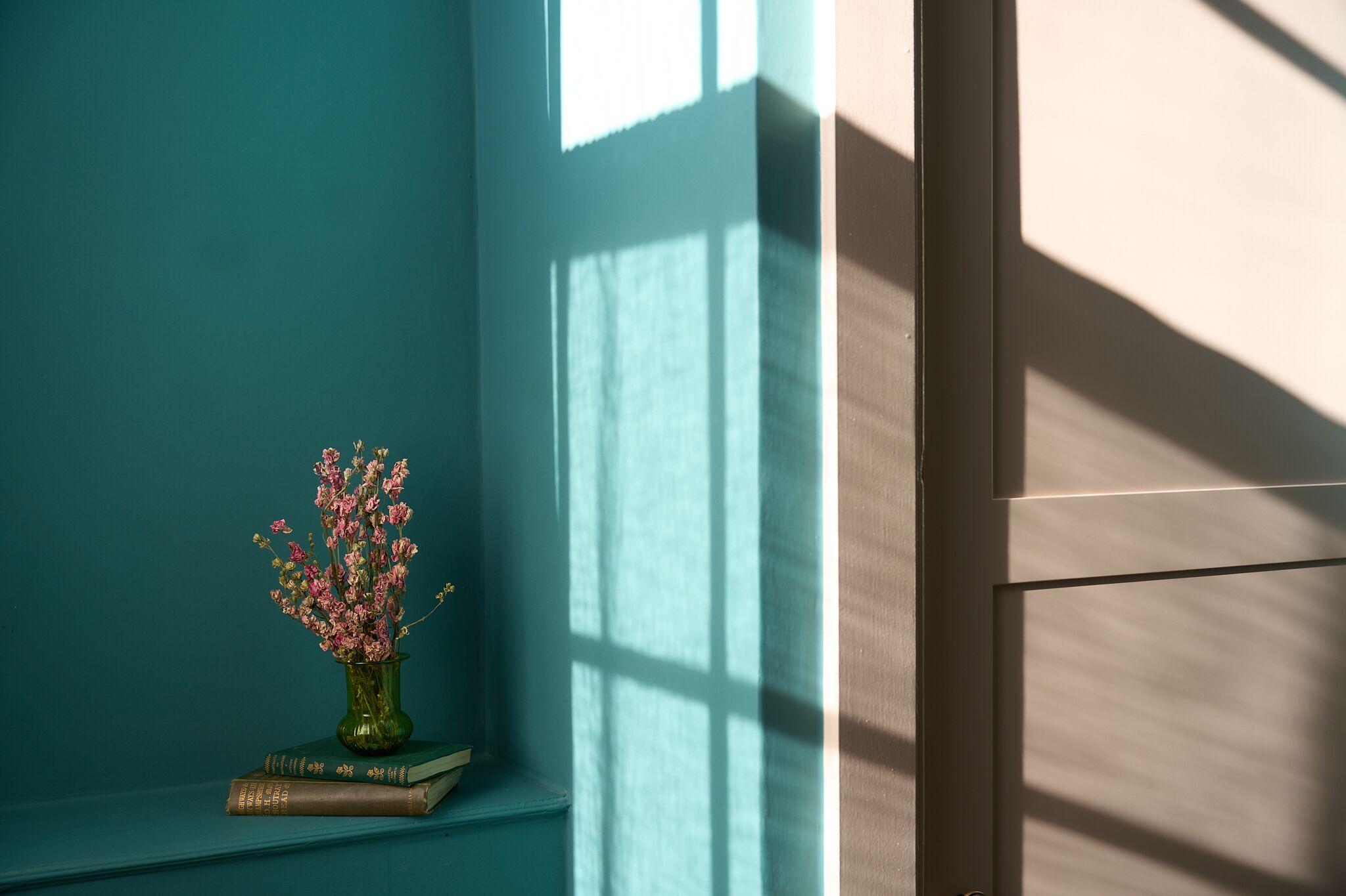A teal wall with a window casting shadows, a green vase with pink flowers, and two books on a ledge.