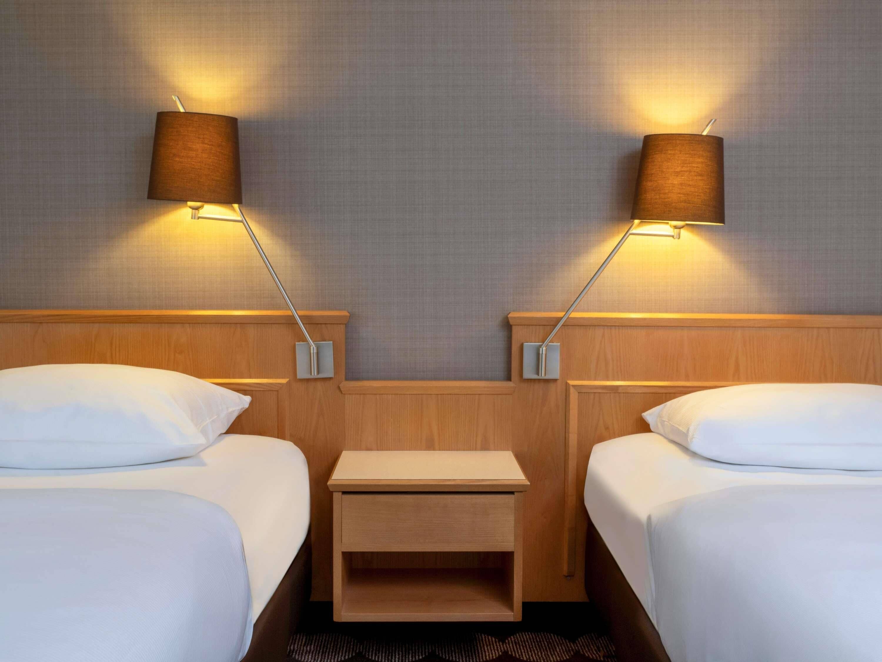 Habitación de hotel con dos camas, una mesita de noche de madera y dos lámparas de pared.