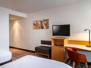Una habitación de hotel con cama, escritorio, televisor, silla y un mueble pequeño.
