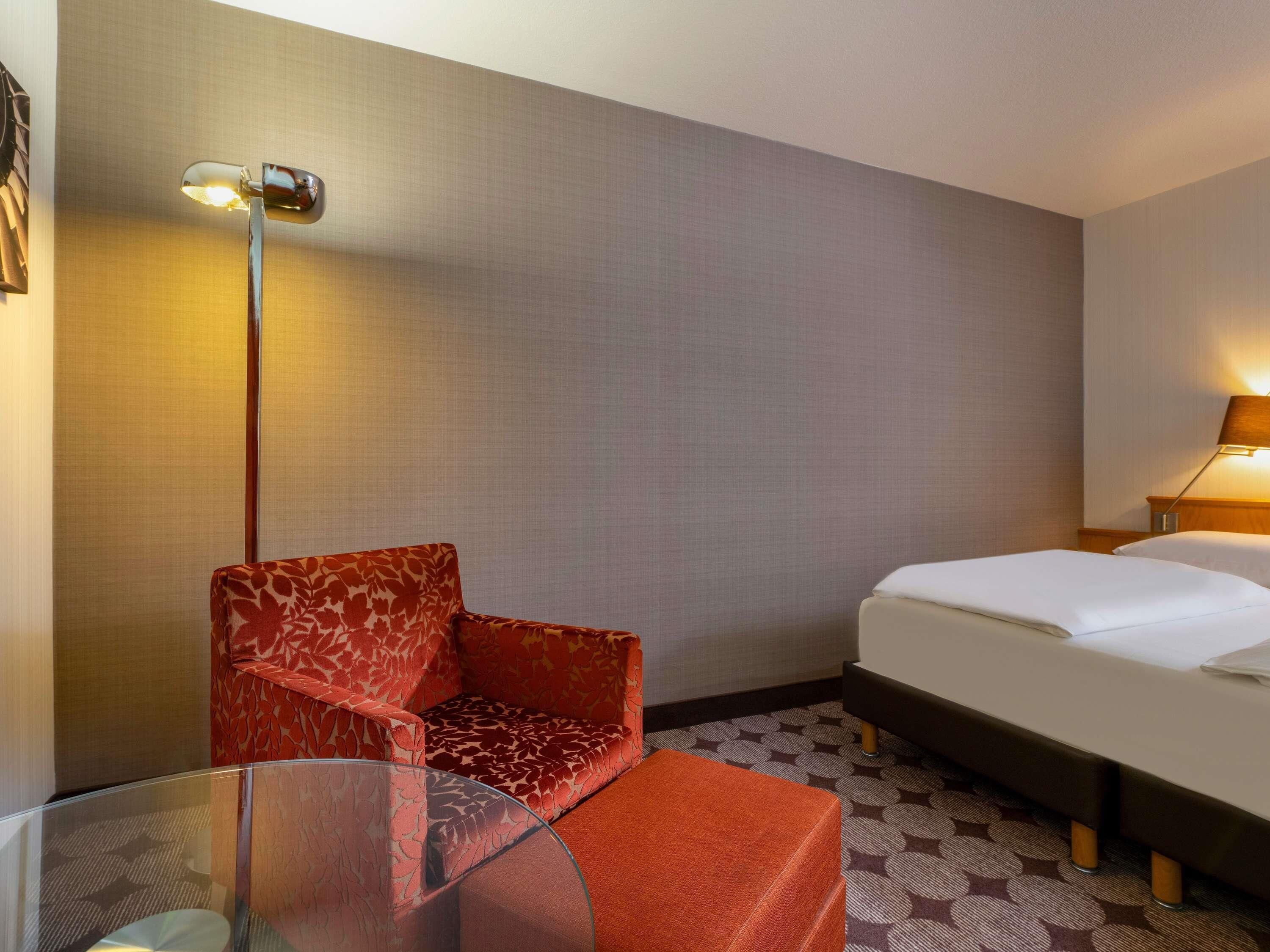 A hotel room with a bed, a red patterned armchair, a glass table, and a floor lamp.