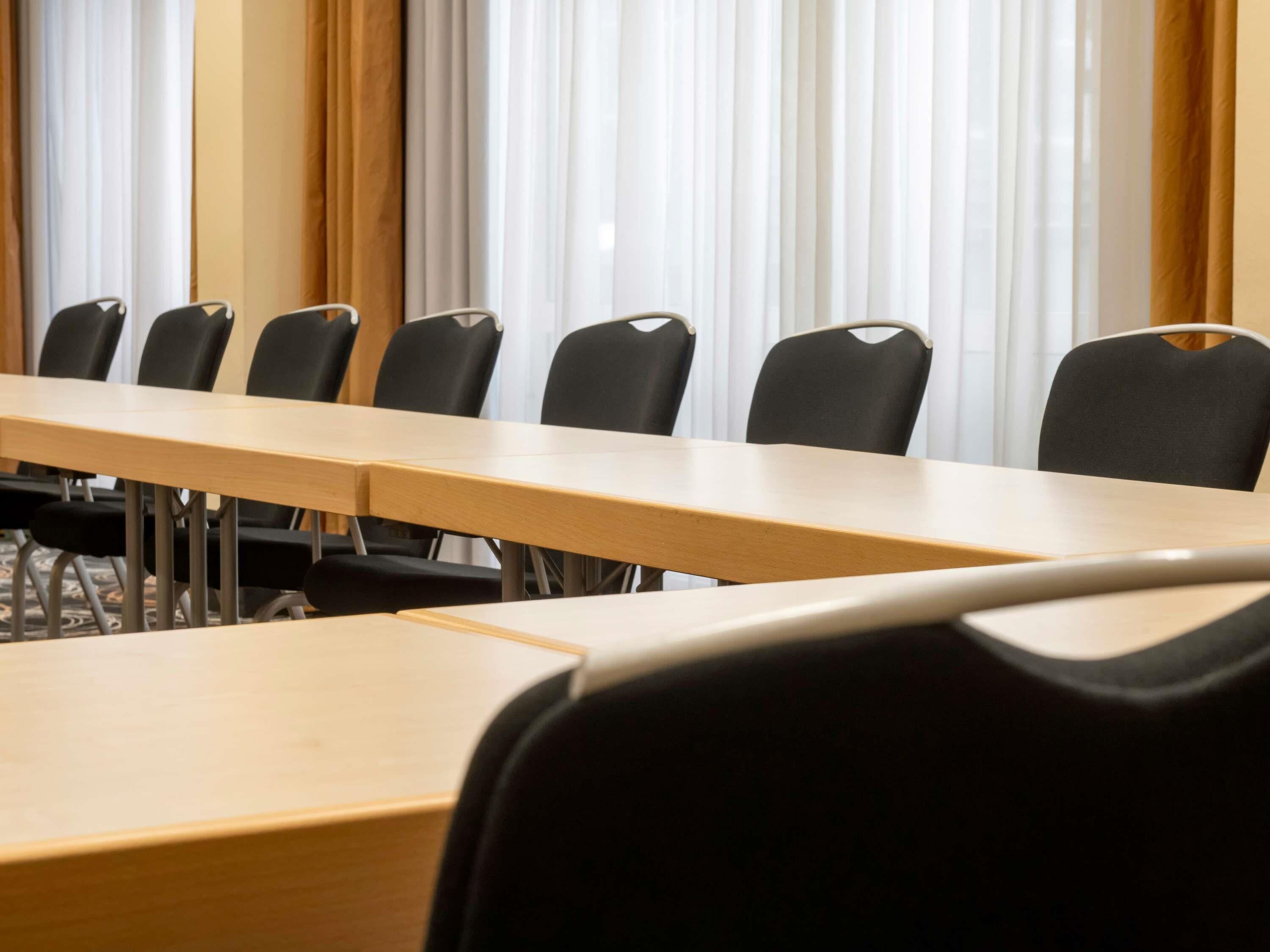 A conference room with a long table and chairs, large windows with curtains, and a carpeted floor.