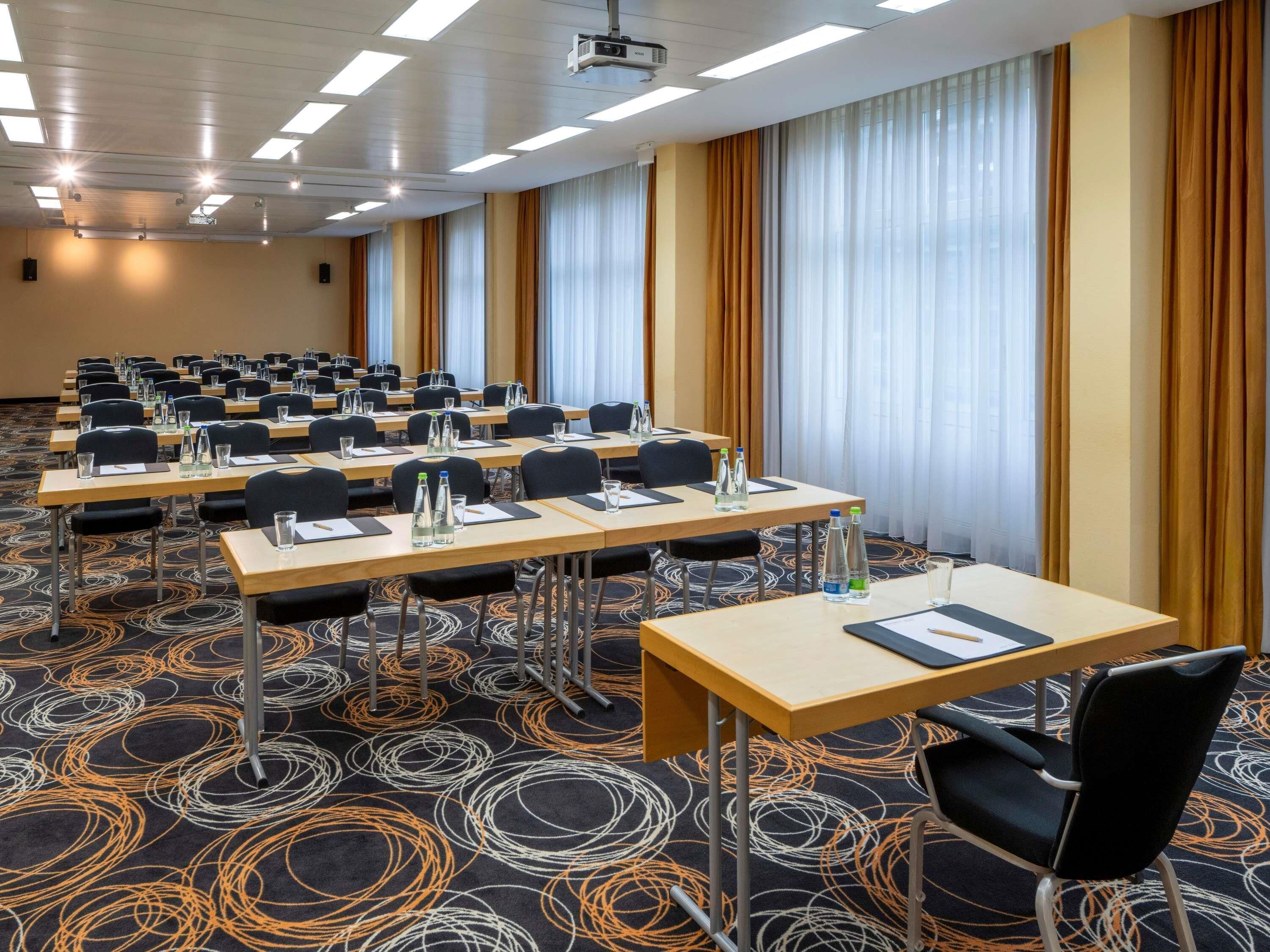 Sala de conferência com mesas, cadeiras e garrafas de água dispostas em fileiras, carpete estampado e amplas janelas com cortinas.