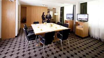 Una habitación de hotel con una mesa de conferencias, sillas, un televisor y un escritorio con lámpara.