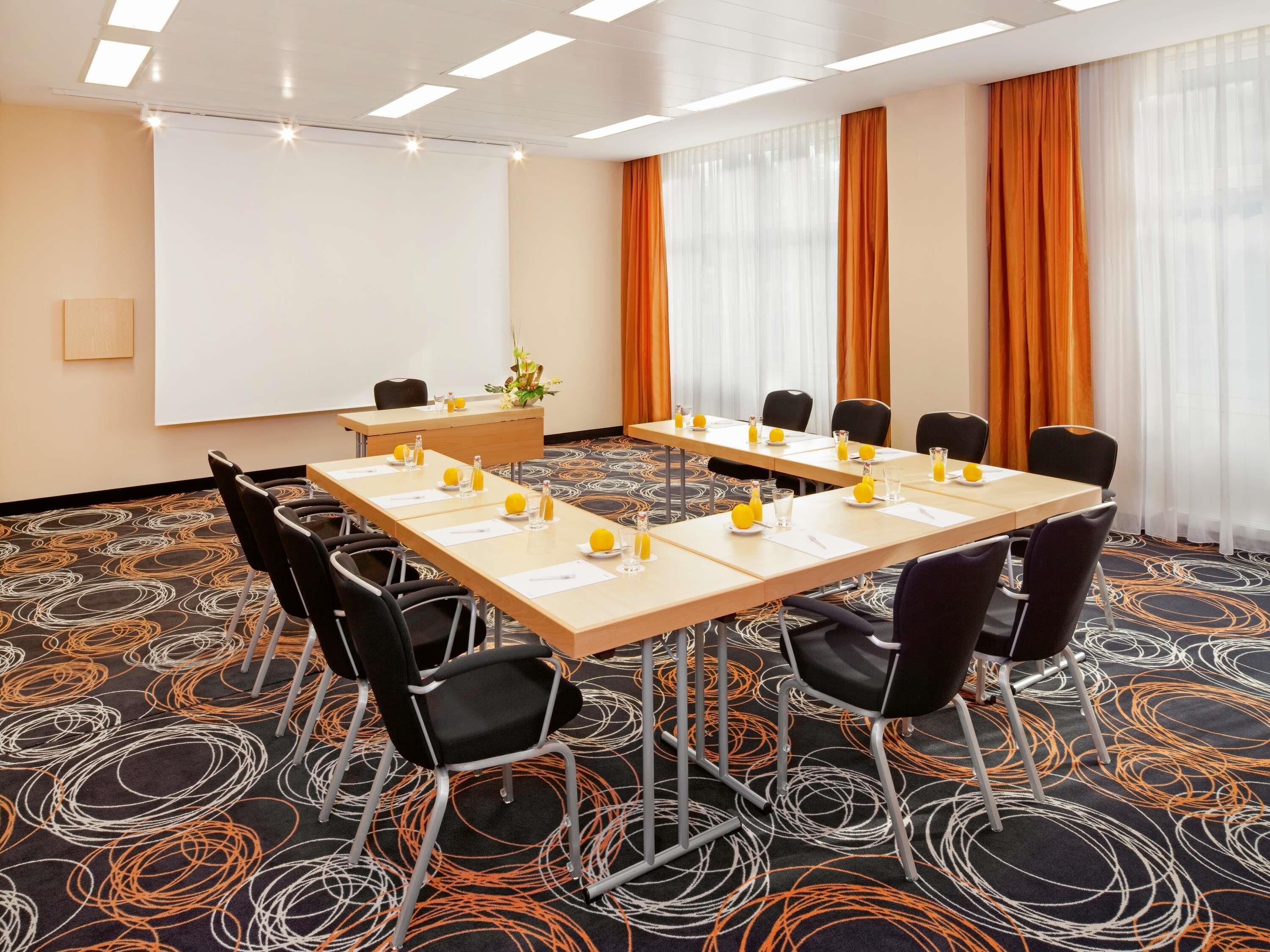 Une salle de conférence équipée d’une longue table, de chaises, d’un écran de projection et d’un tapis orné de motifs circulaires.