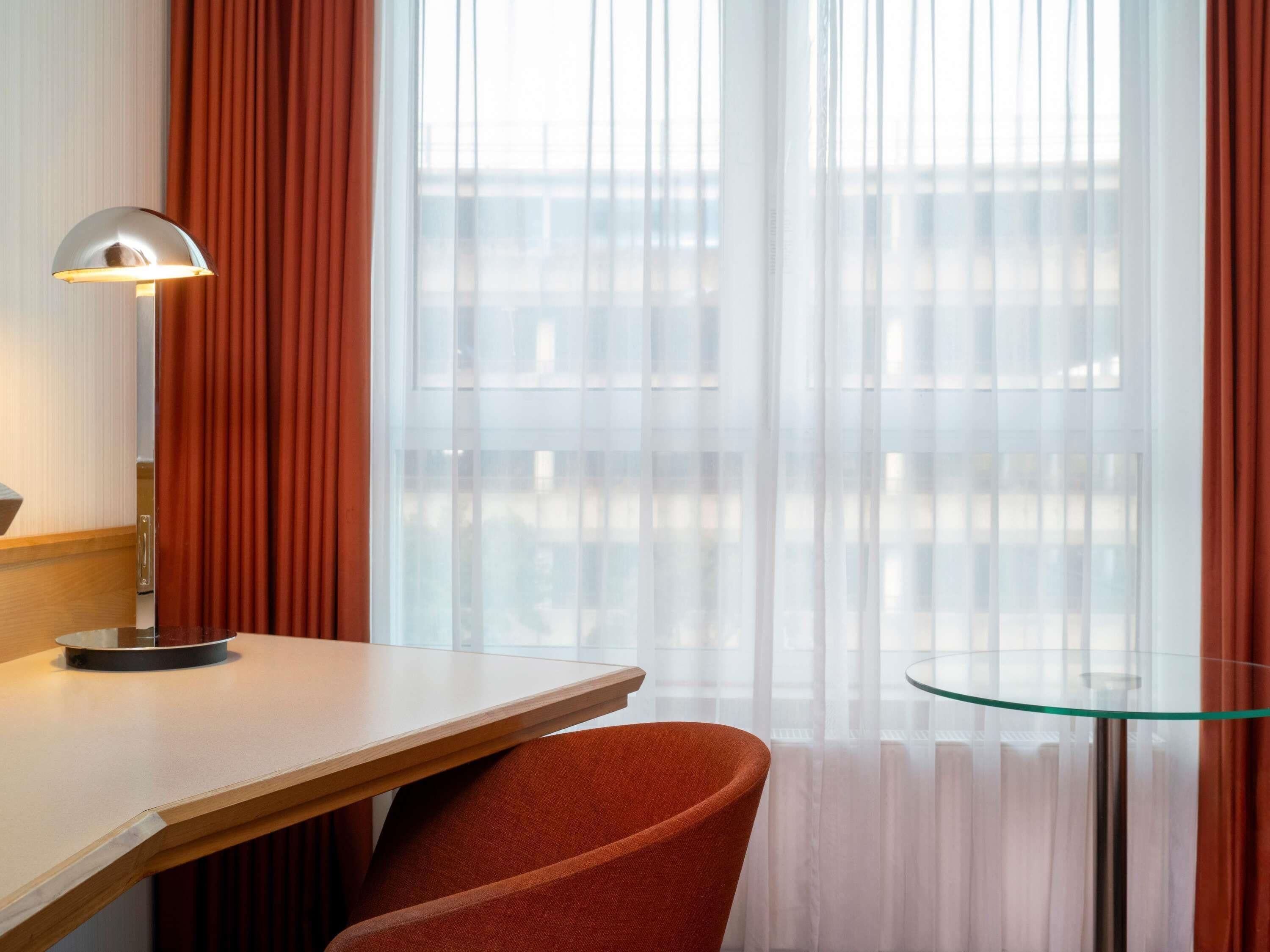 Quarto de hotel com uma escrivaninha de madeira, uma cadeira, uma mesa de vidro e uma janela com cortinas translúcidas.