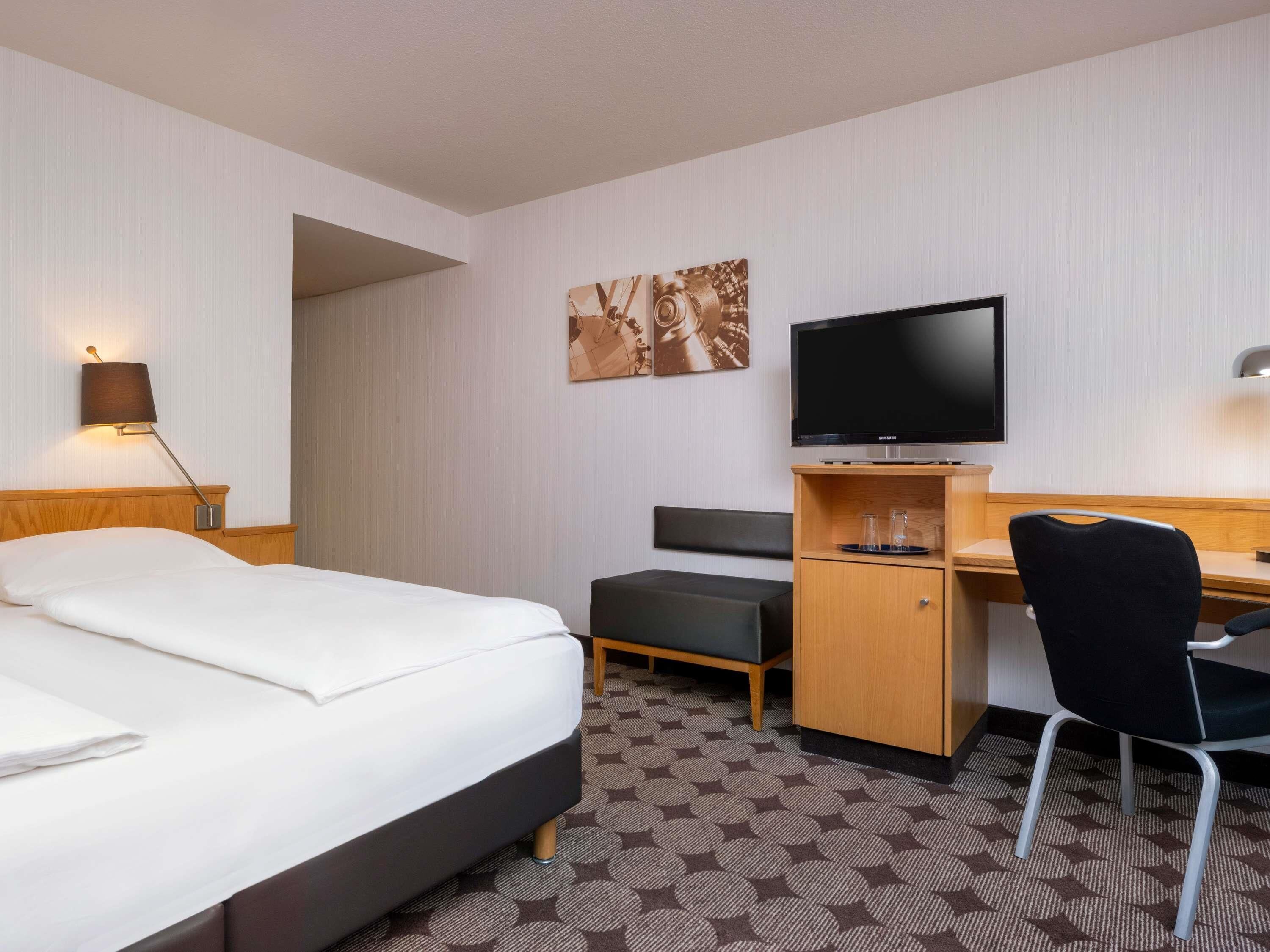 A hotel room with a bed, desk, chair, television, and artwork on the wall.